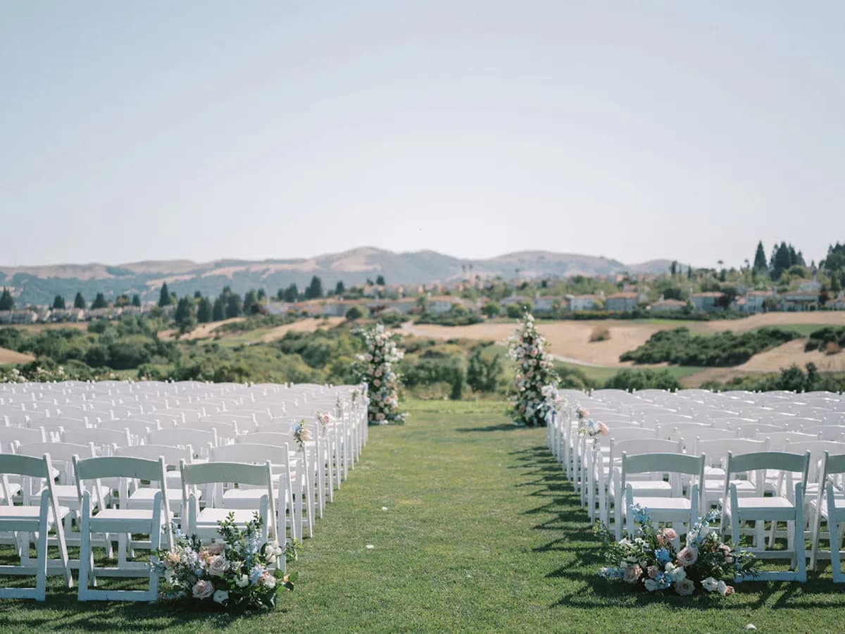 Rows of white chairs lining a grass aisle dressed with pale roses, looking out over rolling hills